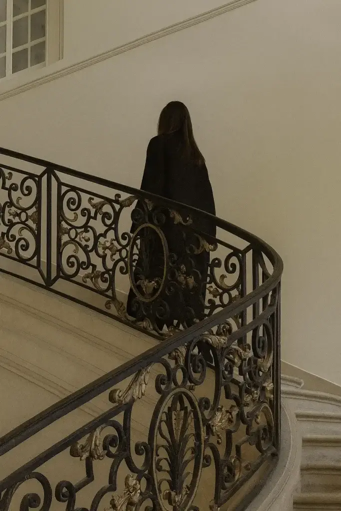 Woman in a long black coat walking up an ornate staircase with wrought iron railing, symbolizing inheritance law and family heritage in Spain.